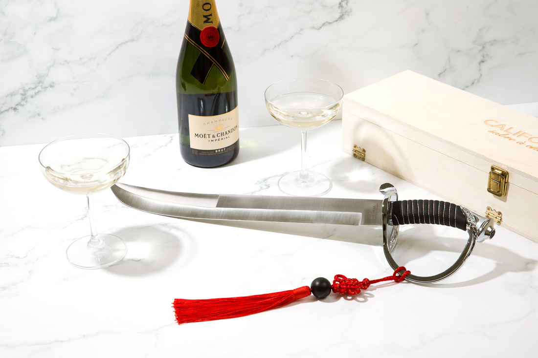 Sword with red tassel on a marble surface next to a bottle of champagne and glasses.
