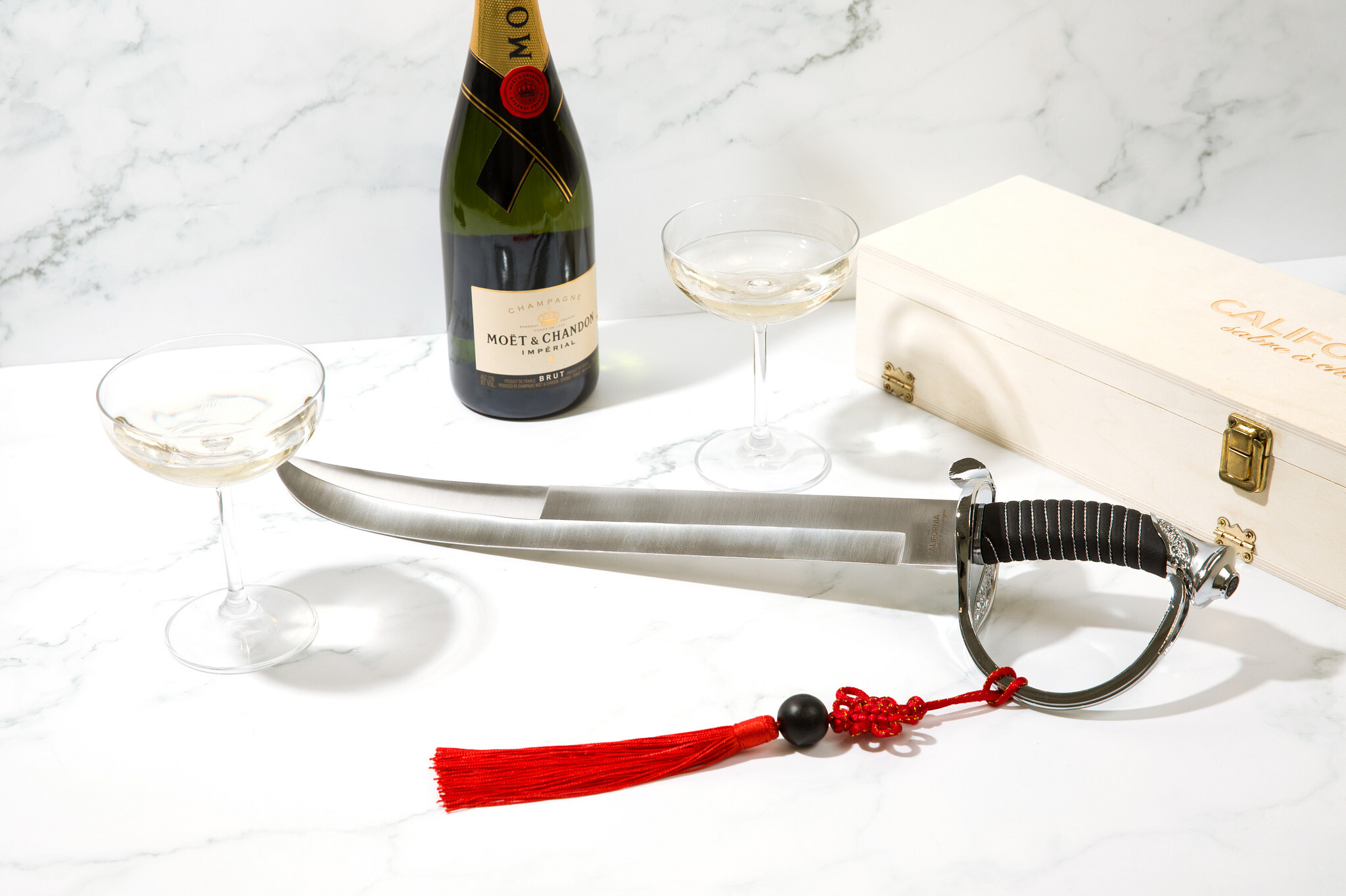 Sword with red tassel on a marble surface next to a bottle of champagne and glasses.